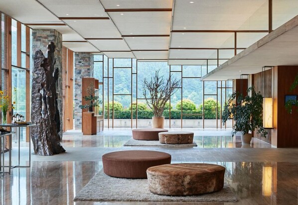 Un lobby espacioso con una gran escultura de piedra, asientos redondos y ventanas amplias que ofrecen vistas a zonas verdes.