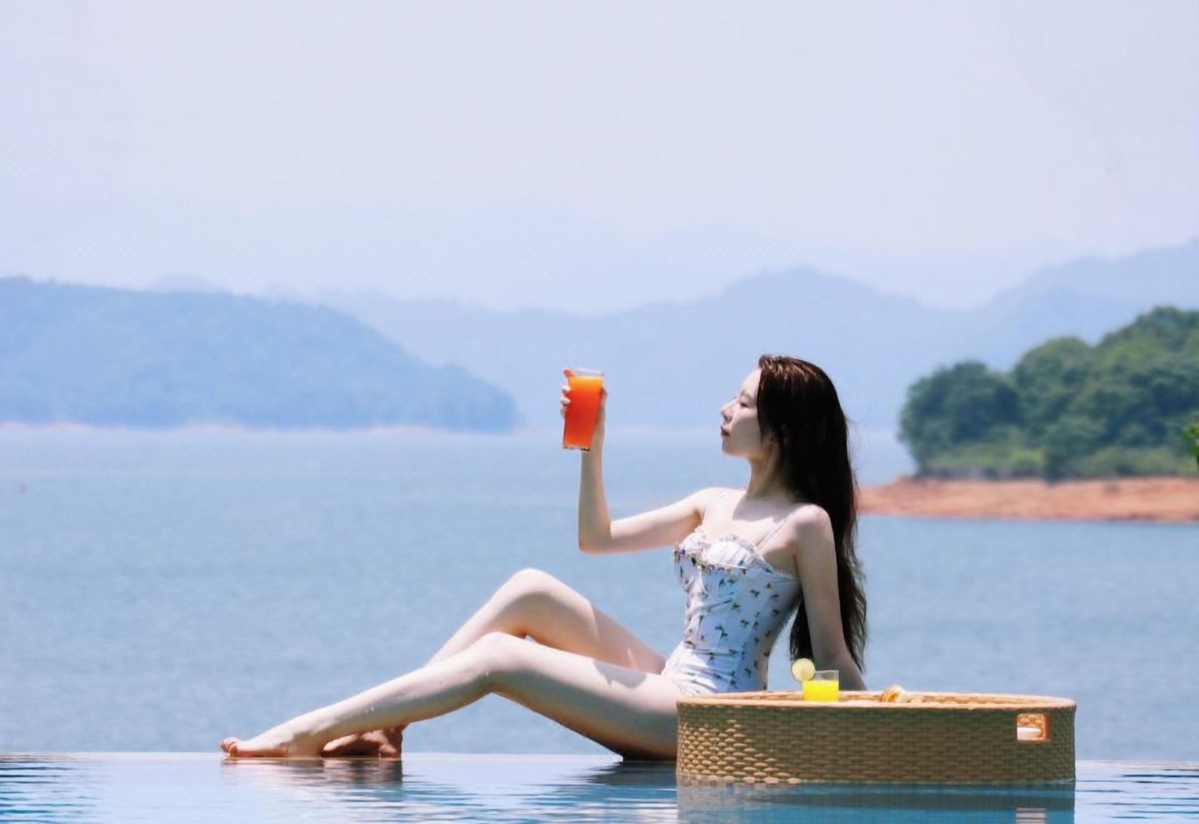 Une personne assise au bord d’une piscine, un verre à la main, avec vue sur une étendue d’eau et des collines lointaines.