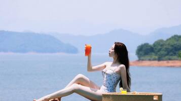 Une personne assise au bord d’une piscine, un verre à la main, avec vue sur une étendue d’eau et des collines lointaines.