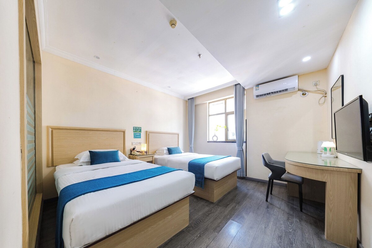Een hotelkamer met twee bedden, een bureau, een stoel, een airconditioning en een raam met gordijnen.