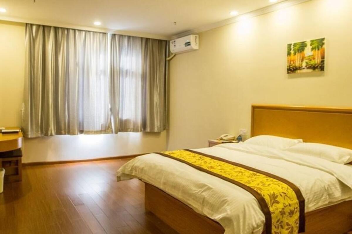 A hotel room with a bed, a desk, a chair, a window with curtains, an air conditioning unit, and a painting on the wall.