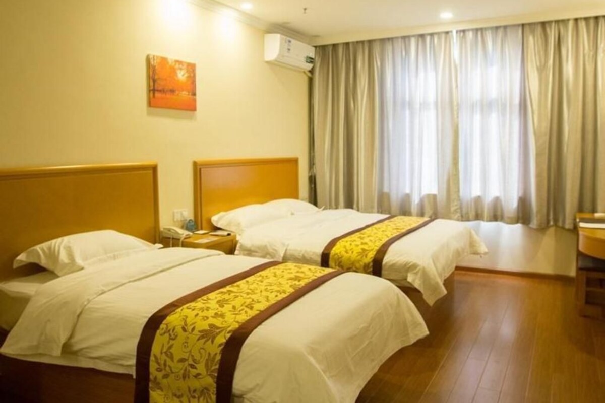 A hotel room with two beds, a bedside table with a lamp, a window with curtains, and an air conditioning unit on the wall.