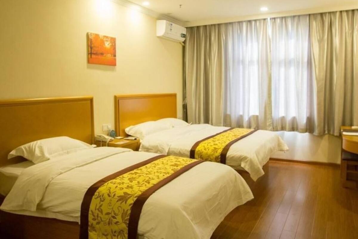 A hotel room with two beds, a bedside table with a lamp, a window with curtains, and an air conditioning unit on the wall.