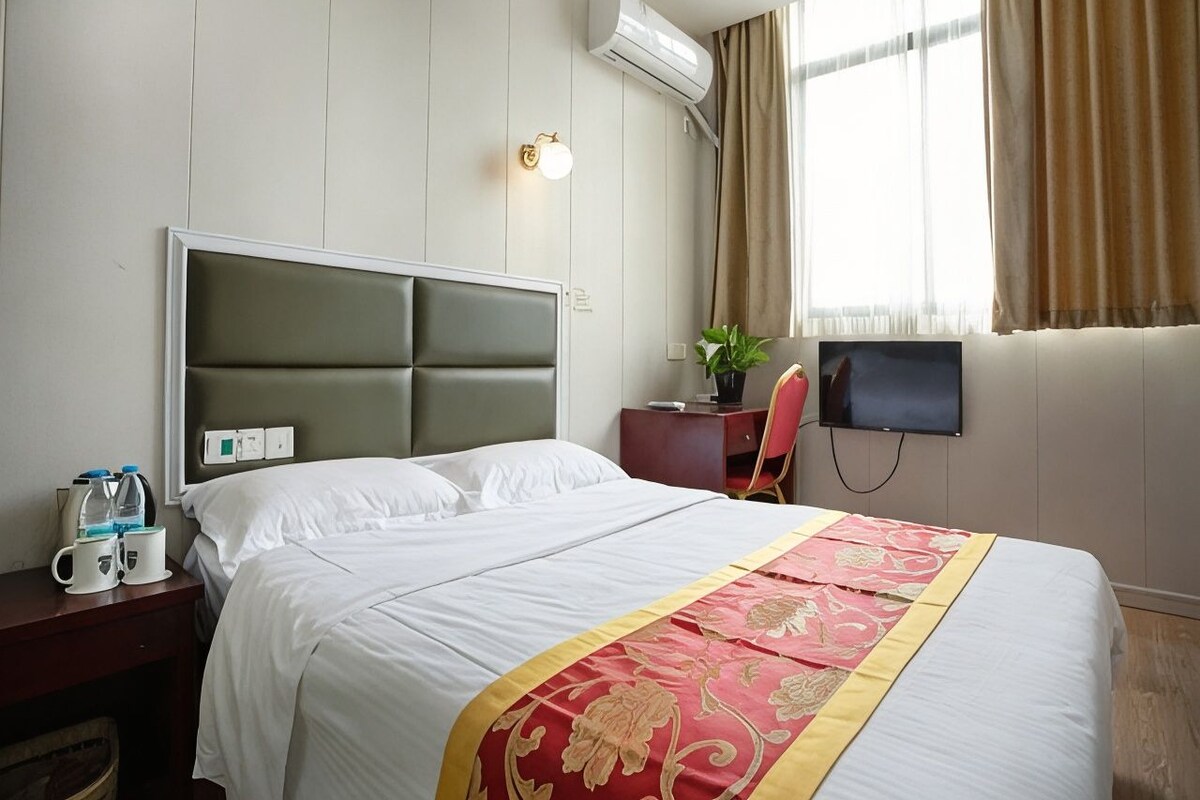 A hotel room with a bed, bedside table, TV, and an air conditioning unit.