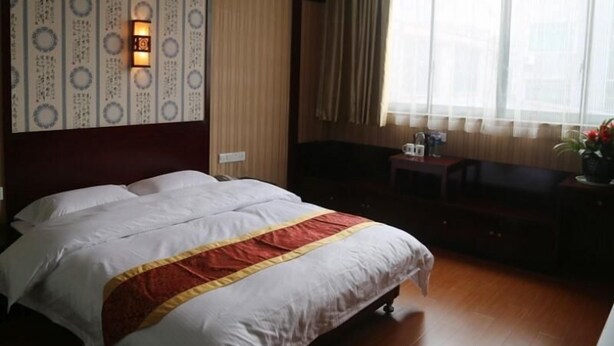 Habitación de hotel con una cama grande, una mesita de noche con lámpara, un pequeño rincón de descanso con una silla y una ventana con cortinas.