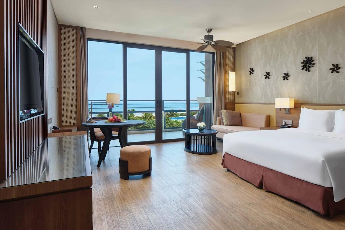 Una habitación de hotel con una cama grande, televisión, mesa de comedor con fruta, sofá y vista al océano.