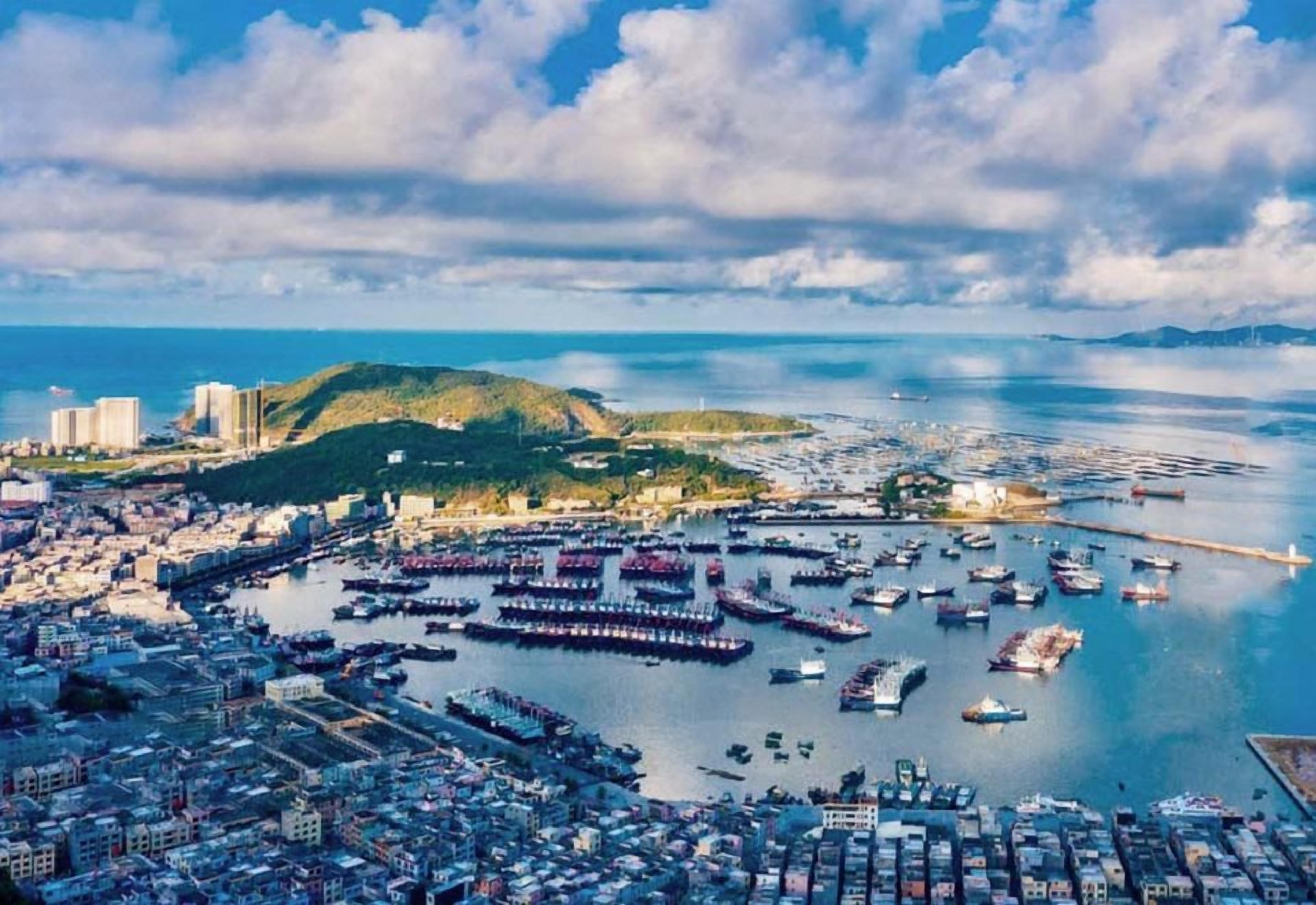 A coastal cityscape with a marina filled with boats, a hilly island in the background, and a dense urban area with buildings.