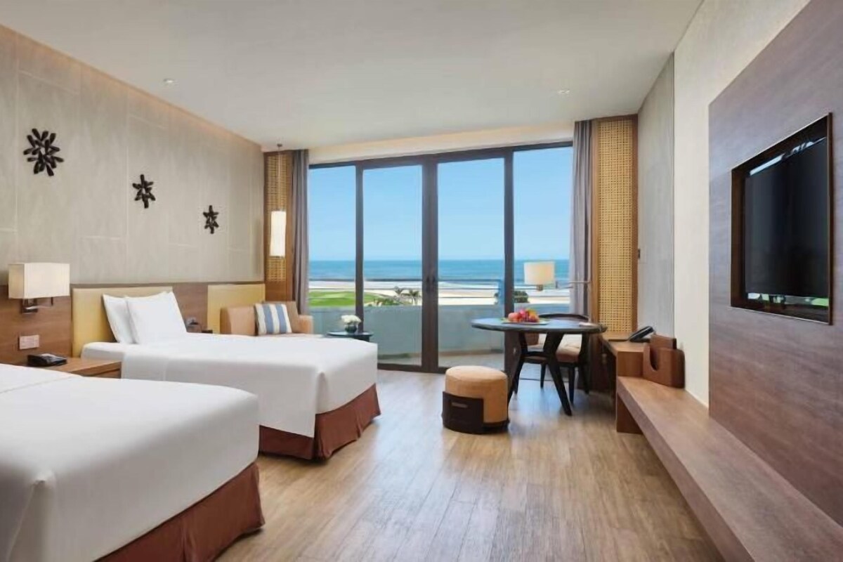 Hotelkamer met twee bedden, een eettafel, een televisie en een balkon met uitzicht op het strand.