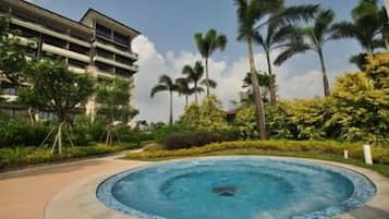 A circular pool with a central fountain, surrounded by lush greenery and palm trees, with a hotel building in the background.