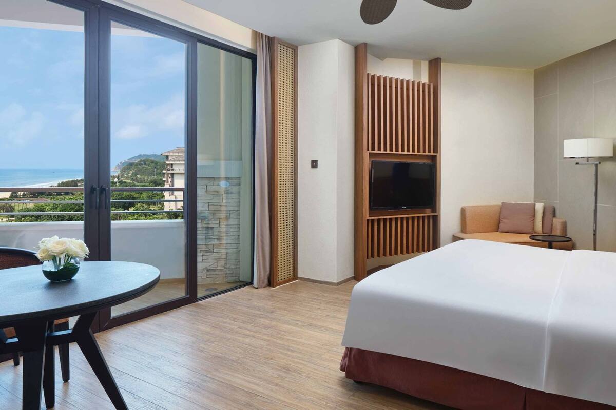 Habitación de hotel con cama, televisor, sofá, mesita y balcón con vistas.