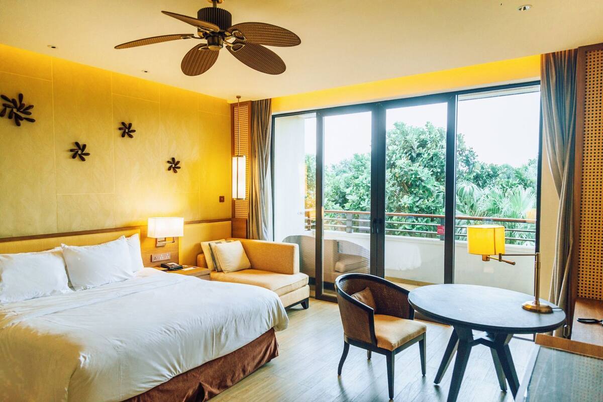 Habitación de hotel con cama, sofá, sillón, mesa y balcón con vista a la vegetación.