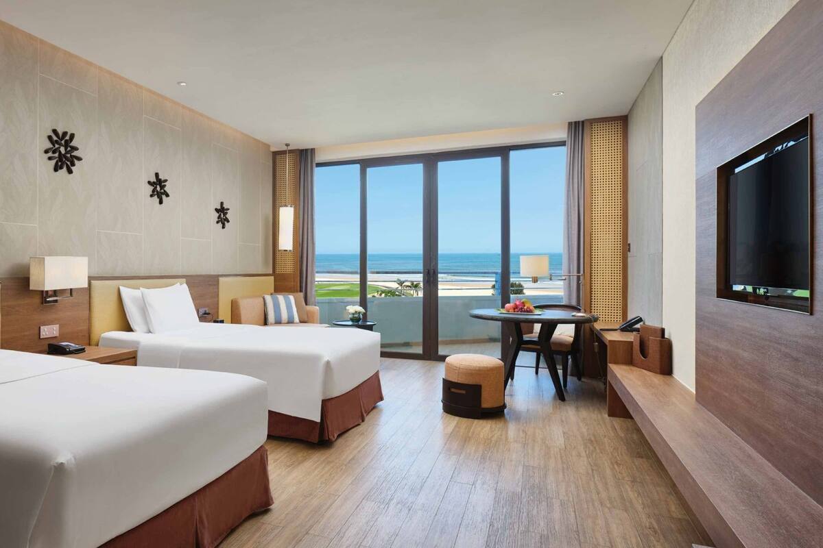 Habitación de hotel con dos camas, zona de comedor, televisor y vista al océano.