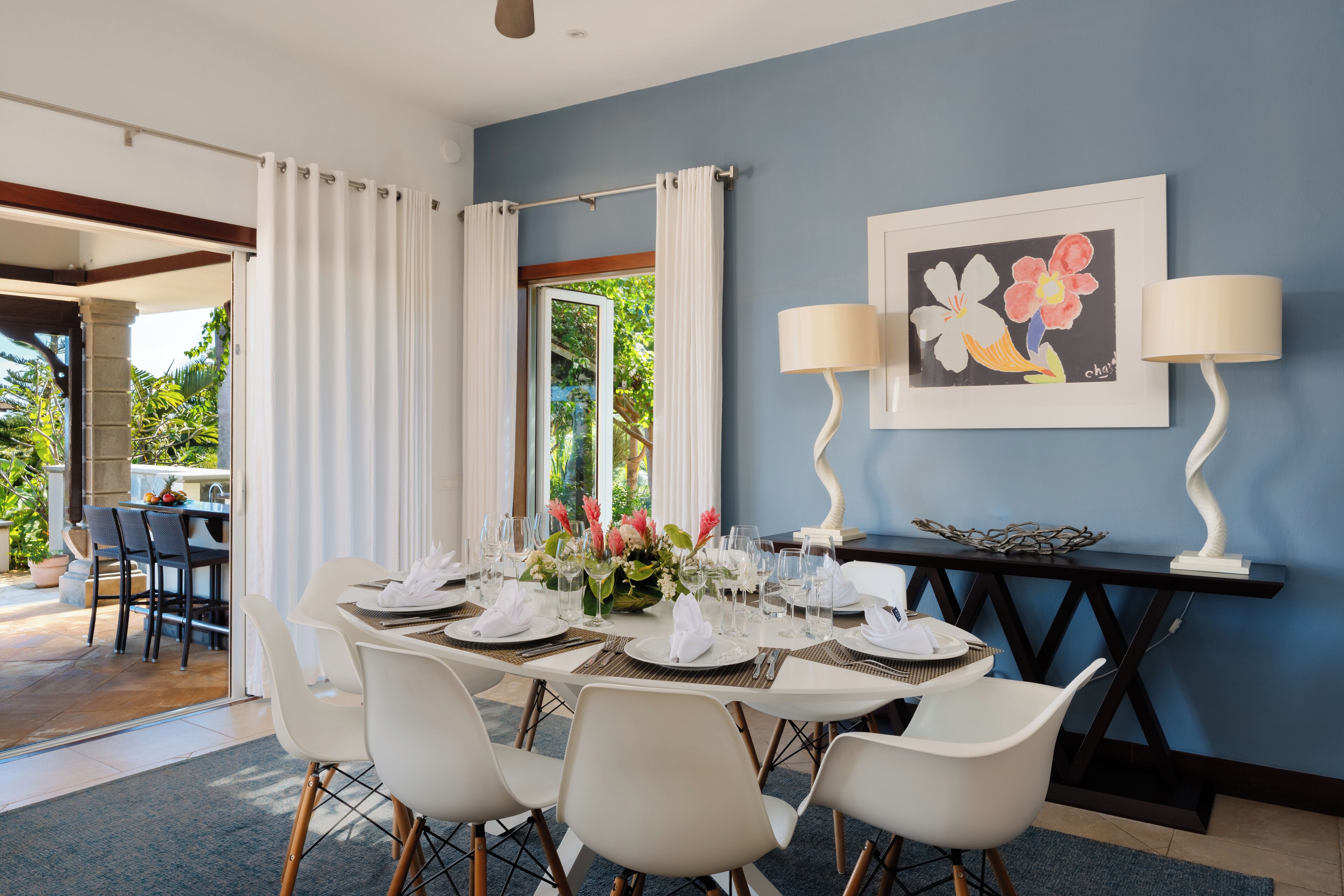 Un comedor con una mesa dispuesta para seis personas, sillas blancas, un cuadro en la pared y una consola con una lámpara.