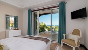 A bedroom with a bed, a chair, a TV, and a sliding glass door leading to a balcony with a view. - Spacious 4-Bedroom Ocean View Villa / Infinity Pool & Outdoor Living (Bel Ombre)