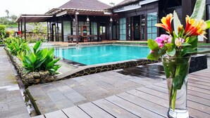 A swimming pool with a wooden deck, a covered seating area, and a building with large windows. - Merlyn Meadows Retreat (Munduk)