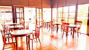 An indoor dining area with multiple wooden tables and chairs, large windows, and a wooden ceiling. - Merlyn Meadows Retreat (Munduk)