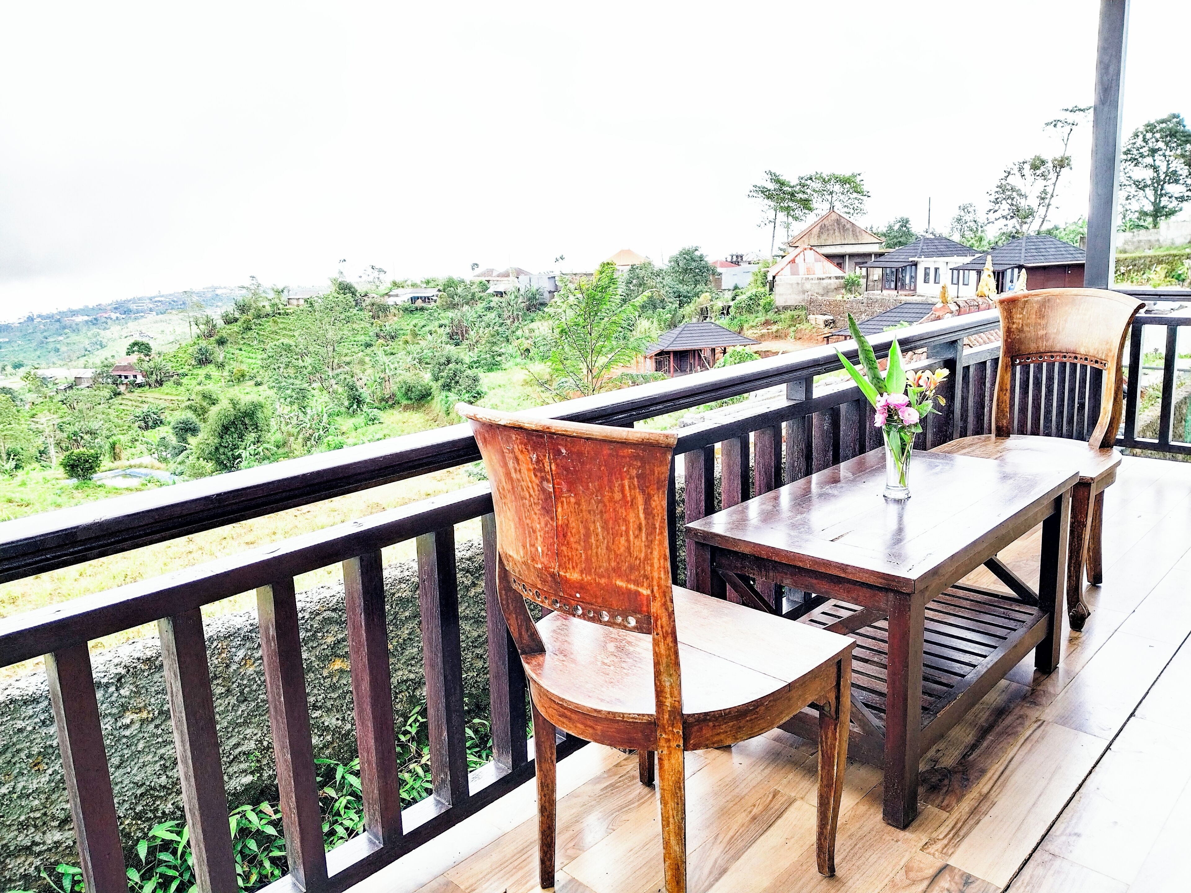 Una terraza de madera con mesa y sillas, con vistas a un paisaje exuberante y casas a lo lejos.