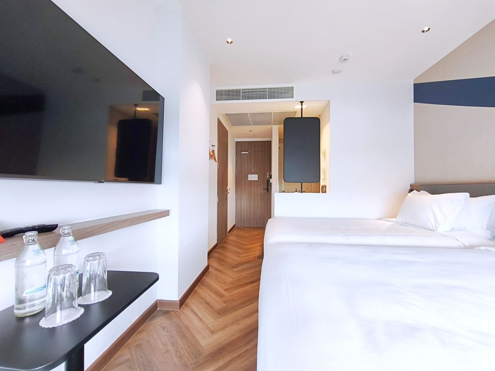 A hotel room with a bed, a TV mounted on the wall, a desk with a bottle and glasses, and a hallway leading to a door.