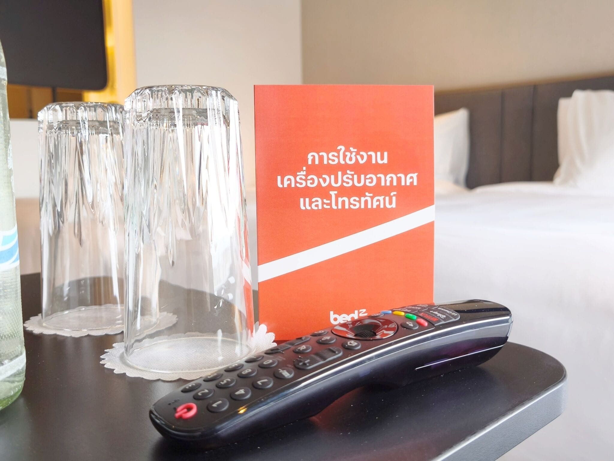 Um controle remoto, dois copos vazios e uma placa vermelha com texto tailandês sobre a mesa de um quarto de hotel.