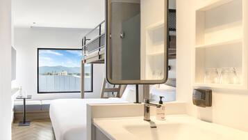 A modern hotel room with a bunk bed, a large window with a city view, a sink with a soap dispenser, and a small table with a bottle.