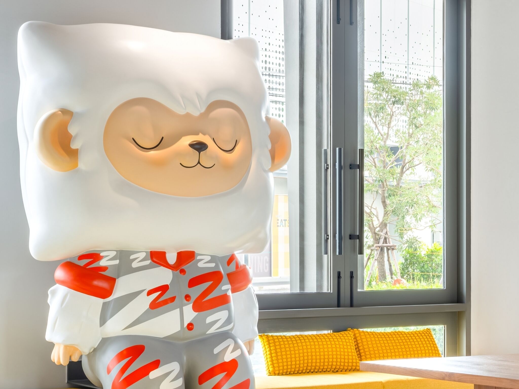 A plush toy with a bear-like face and a sleeping expression, positioned in front of a window with a view of greenery and buildings.