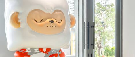A plush toy with a bear-like face and a sleeping expression, positioned in front of a window with a view of greenery and buildings.