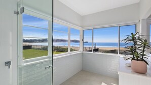 Quarto de canto com grandes janelas com vista para uma praia, um vaso de planta e uma parede com azulejos brancos.