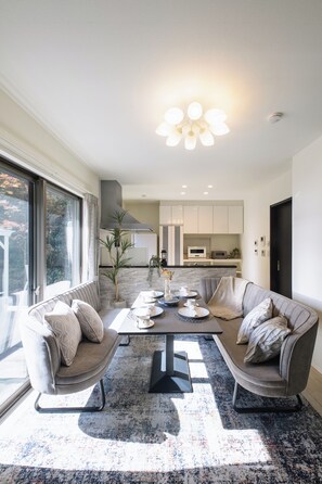 Een moderne woonkamer met een zithoek, een salontafel en een eettafel met stoelen. Achtergrond is een keuken met witte kastjes en een magnetron.