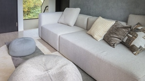 Un salon moderne avec un grand canapé d’angle, un pouf rond, une œuvre d’art abstraite accrochée au mur et une vue sur les montagnes par une grande fenêtre.
