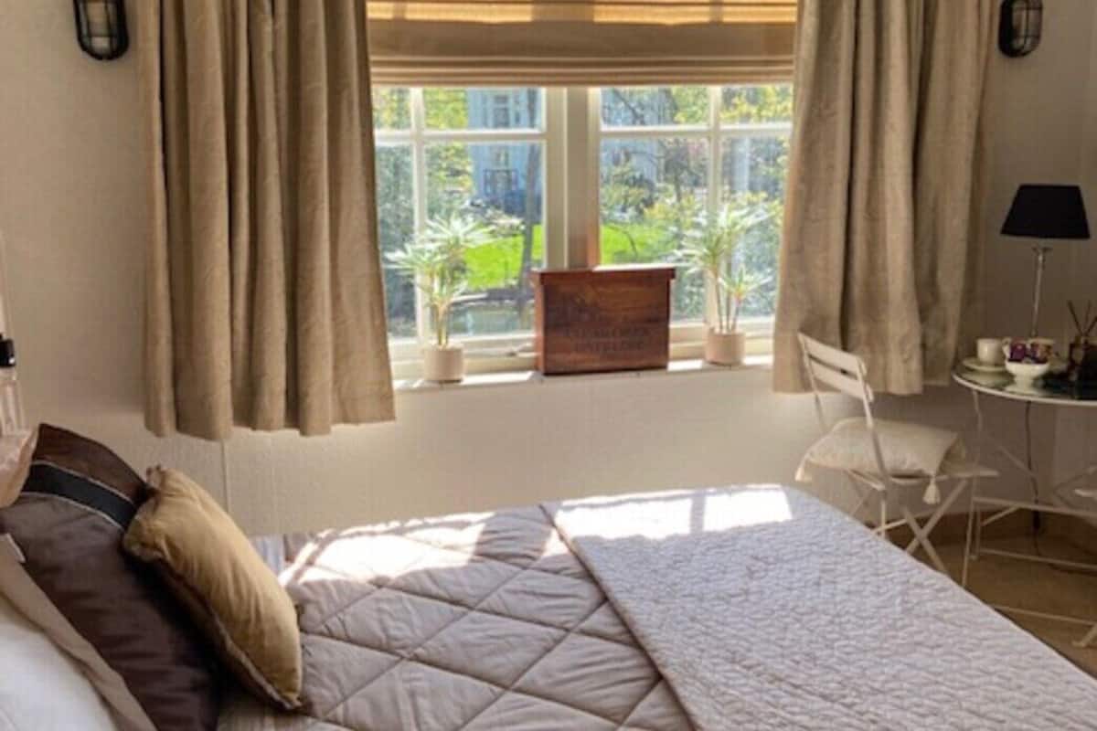 A bedroom with a bed, curtains, a window with a view, a small table with a lamp, and a chair.