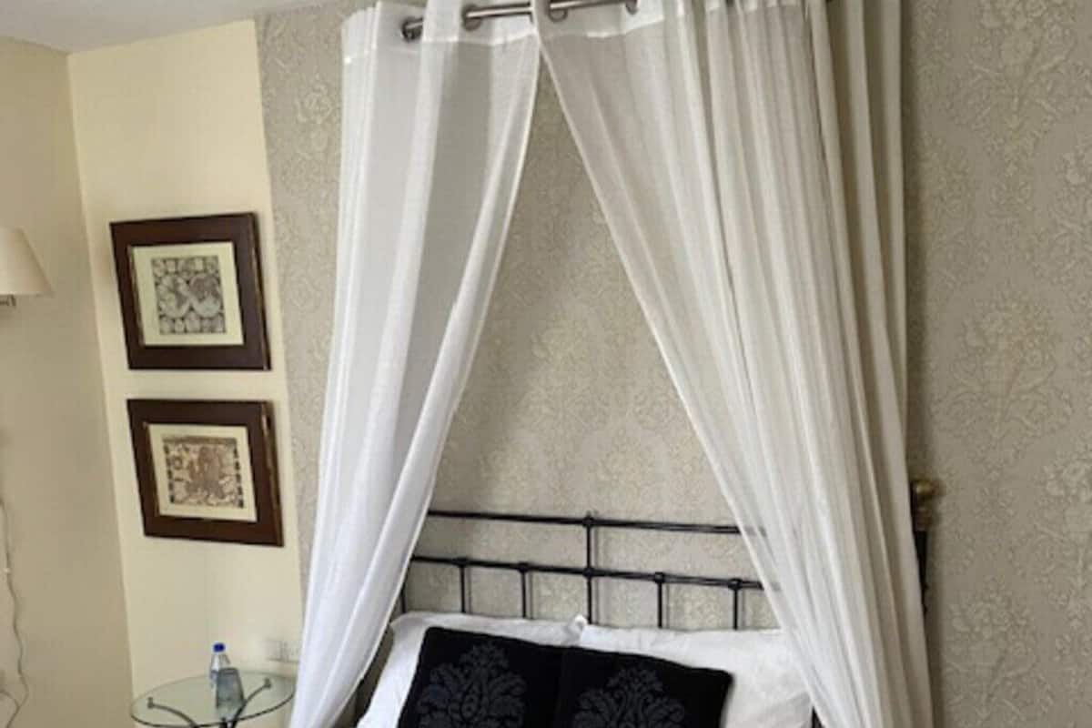 A bed with a canopy, framed pictures on the wall, and a small table with a bottle.