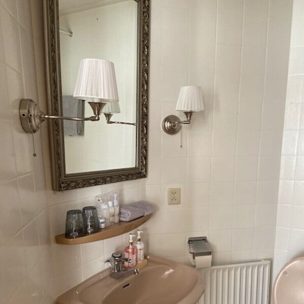 Un baño con espejo, dos lámparas de pared, lavamanos con artículos de aseo y un radiador.
