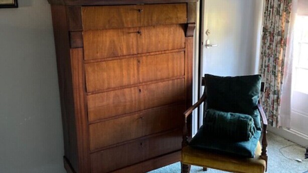 Un mueble de madera con cajones, una silla con cojín, una lámpara y un cuadro en la pared.