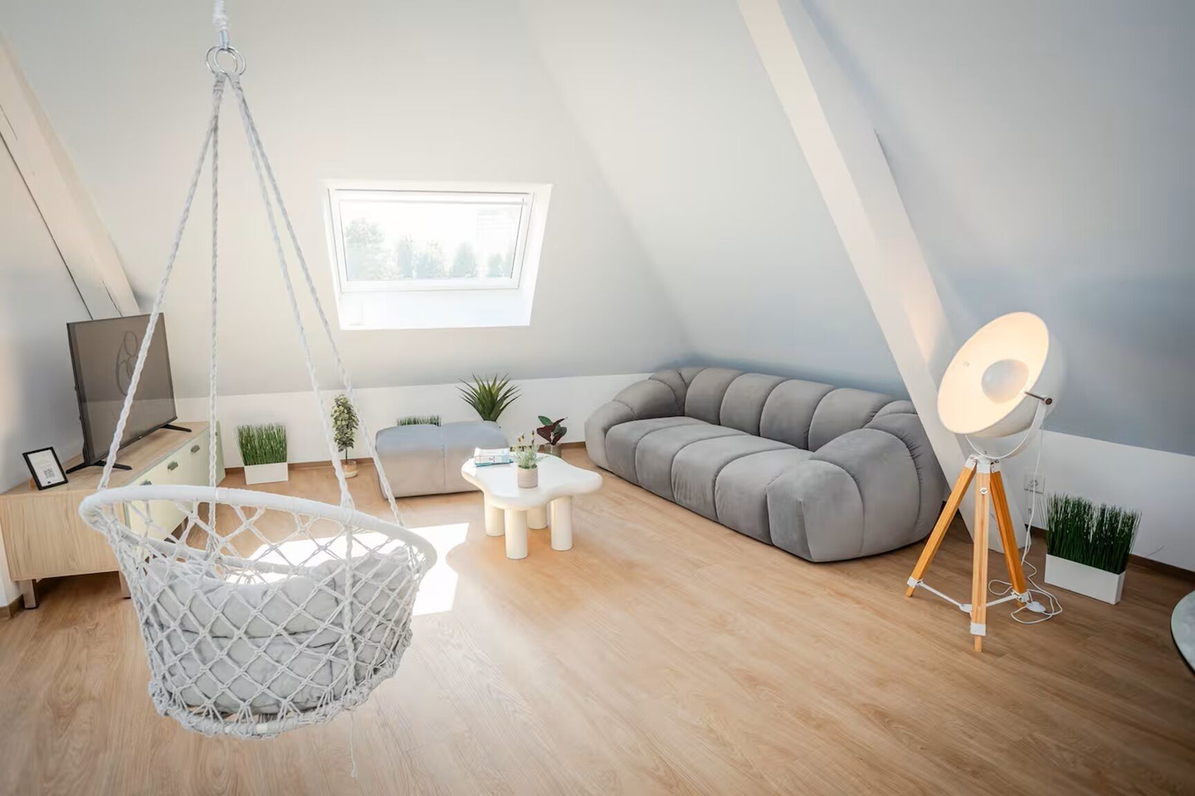 Ein Loft-Zimmer mit Hängekorbsessel, Sofa, kleinem Tisch, Stehlampe und Fenster mit Aussicht.