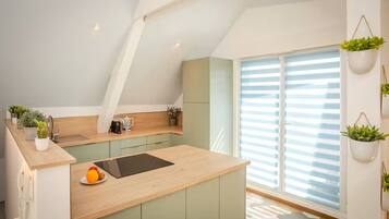 Une cuisine moderne avec un plan de travail en bois, des meubles de couleur verte, une plaque de cuisson à induction noire et une fenêtre avec des stores.