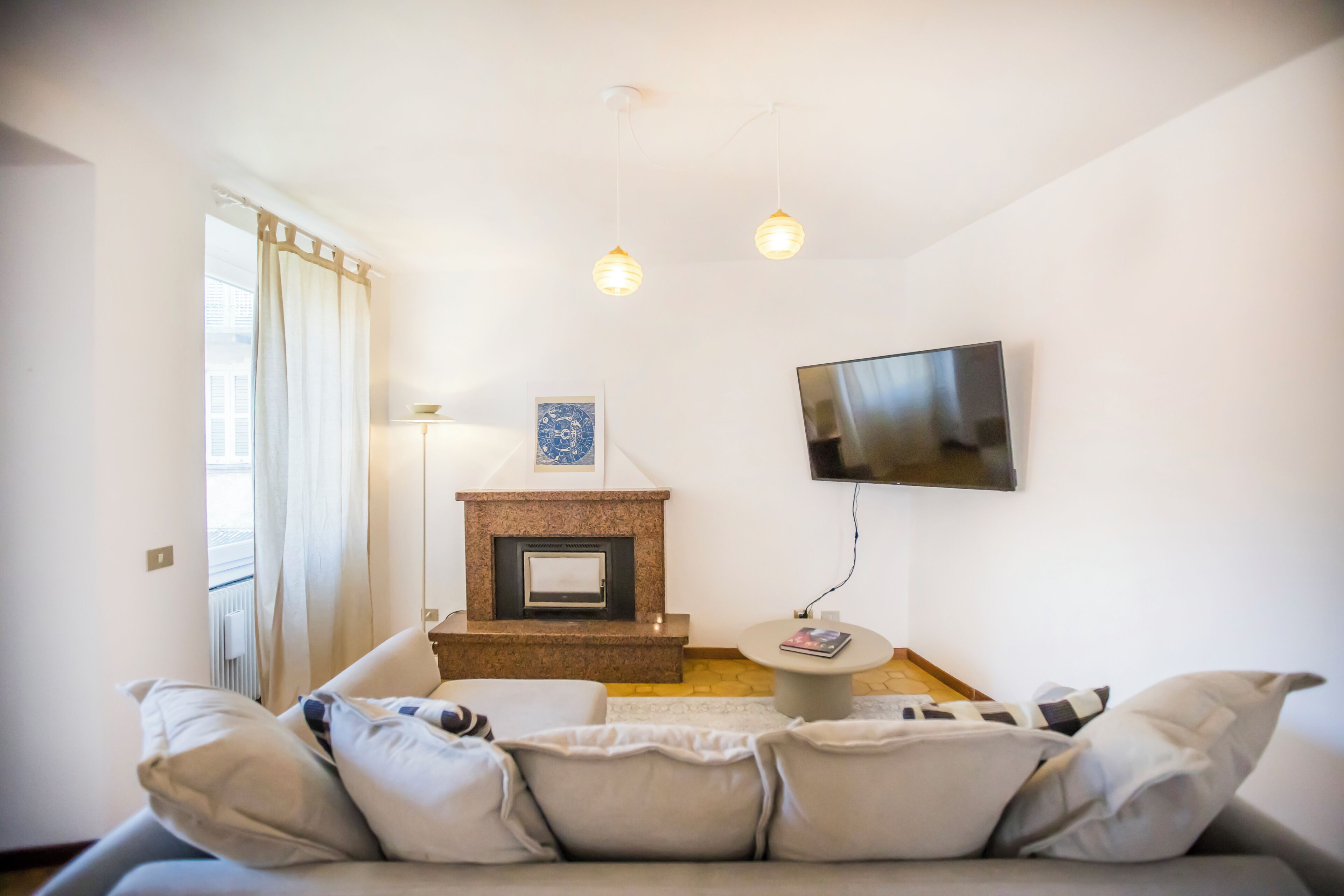 A living room with a fireplace, a sofa, a TV mounted on the wall, and a window with curtains.