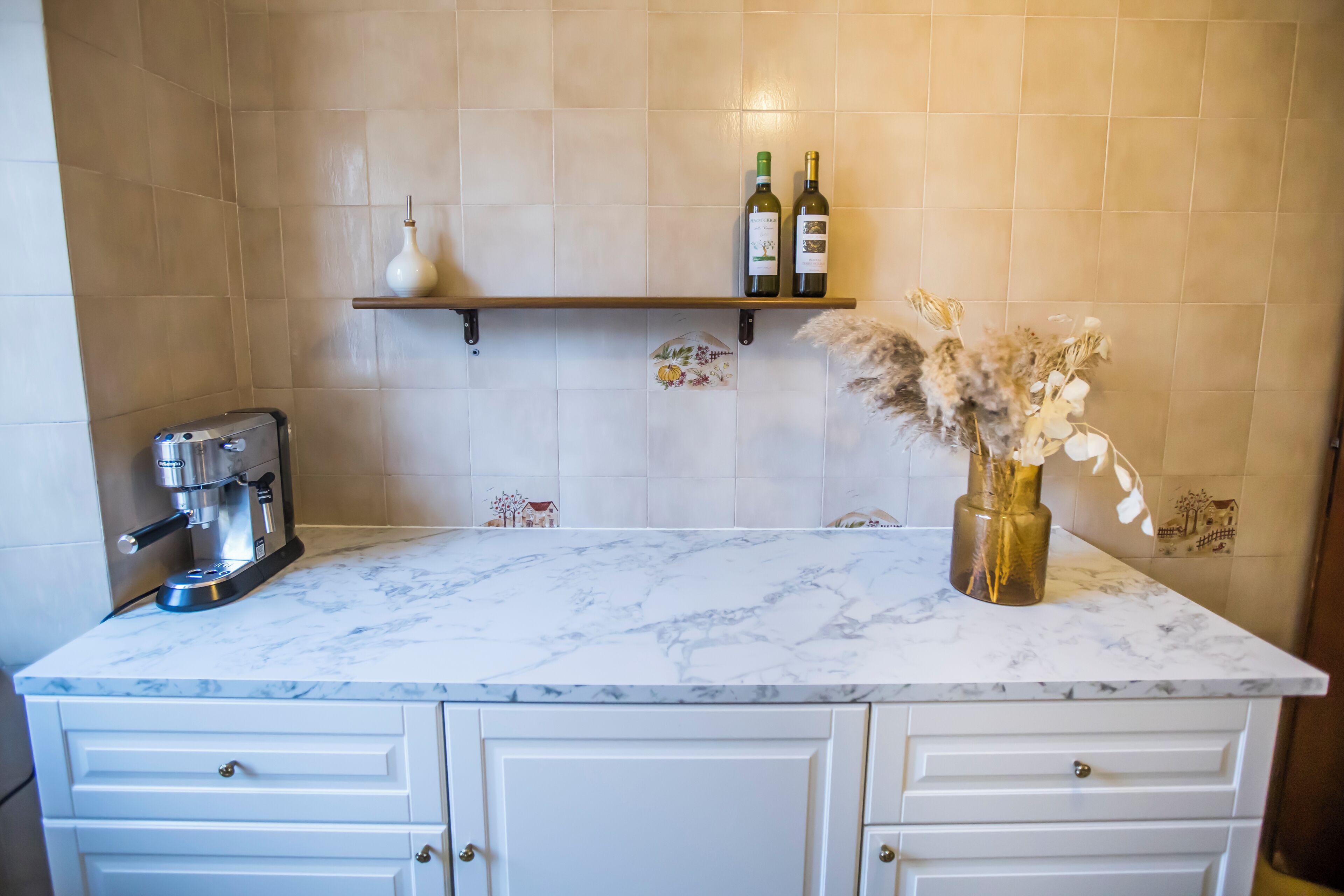 A kitchen with a marble countertop, a coffee machine, a shelf with bottles, and a vase with dried flowers.