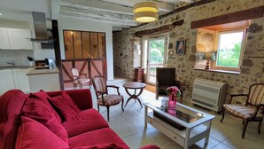 A cozy living room with a red sofa, a coffee table, and striped chairs. A kitchen with white cabinets and a stone wall is visible in the background. - Maison en pierre avec piscine – Nature & calme en Vallée de la Dordogne (Bilhac)