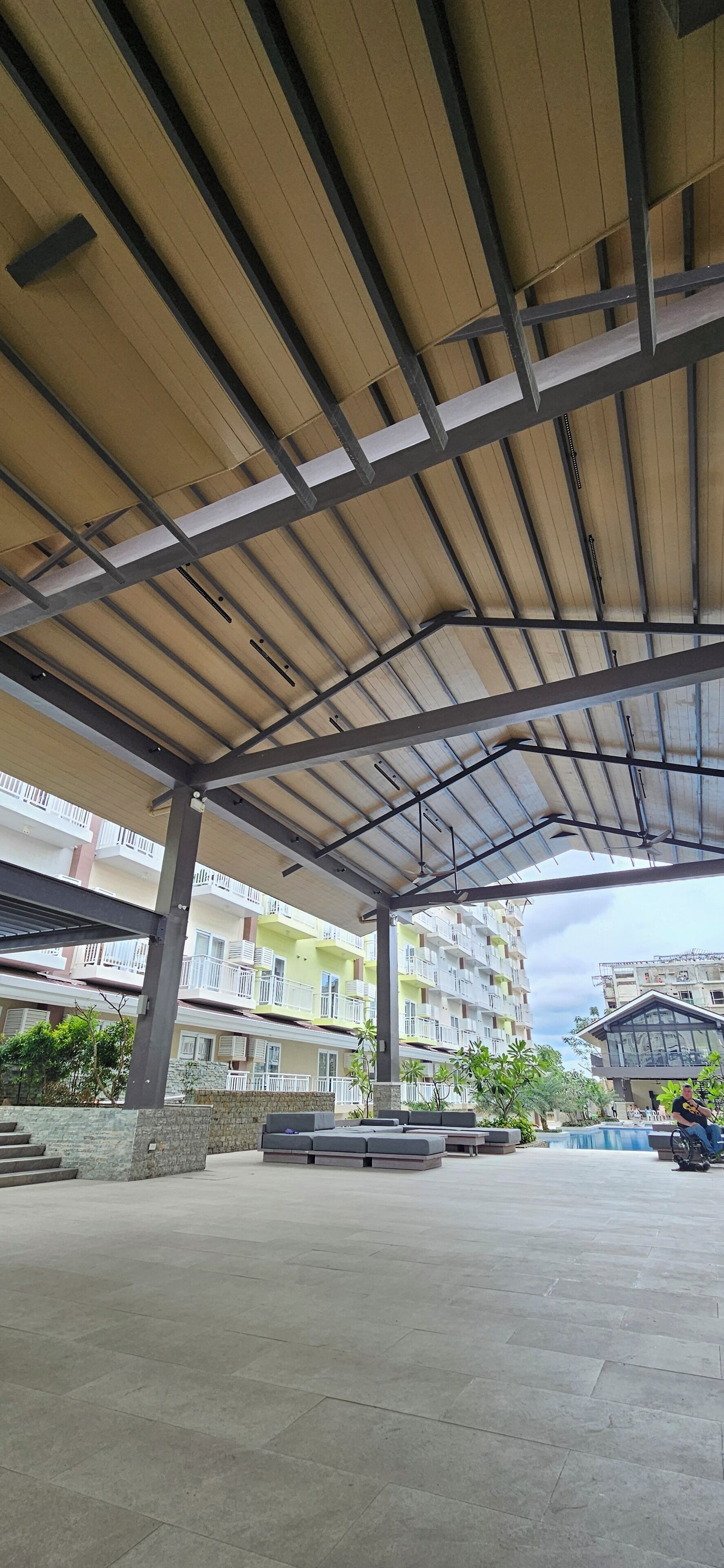 Un espacio al aire libre cubierto con suelo de baldosas, vigas metálicas y vista a edificios de varios pisos al fondo.