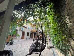 A hanging chair in a courtyard with a brick building and potted plants in the background. - Fayz (Samarkand)