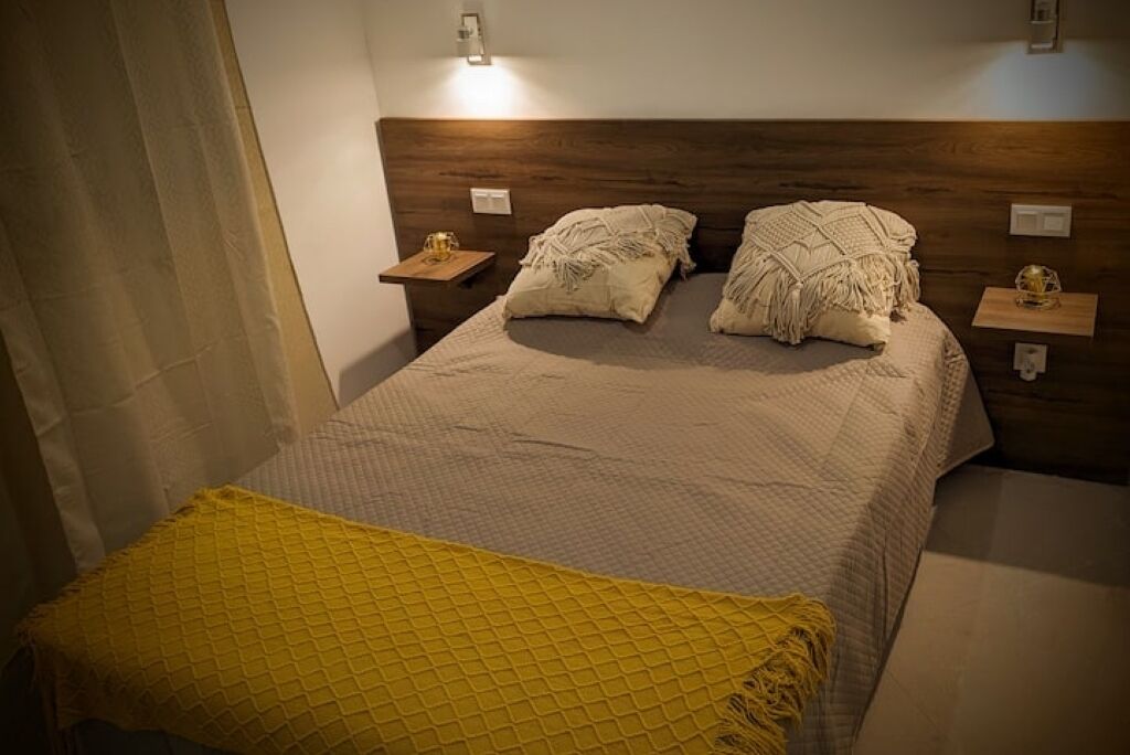 A bedroom with a bed, two bedside tables, a nightstand, a yellow throw blanket, and a wall-mounted light fixture.