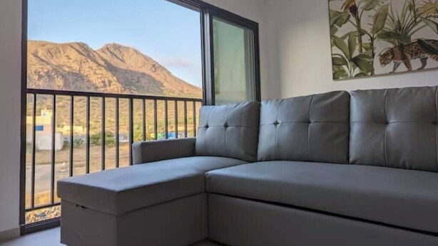 Een moderne woonkamer met een grijze modulaire bank, een groot raam met uitzicht op bergen, en een kunstwerk met een tropische plant aan de muur.