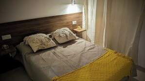 Een bed met een houten hoofdbord, twee kussens, een gele plaid, een nachtkastje met een lamp en een gordijn.