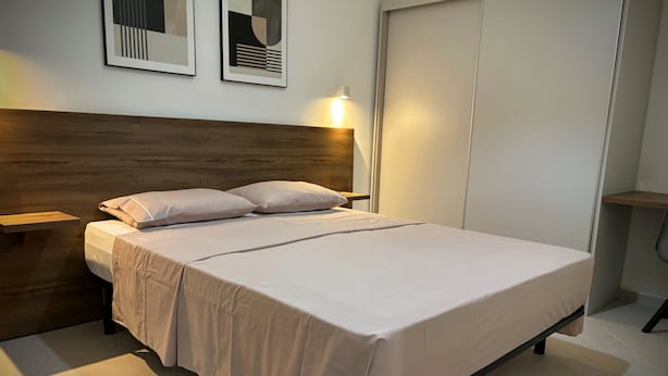 Una cama bien hecha con cabecera de madera, una mesita de noche con una lámpara y dos obras de arte abstracto enmarcadas en la pared.