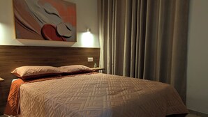 A neatly made bed with a patterned quilt, a bedside table with a lamp, a wall-mounted artwork, and a window with curtains. - Tarrafal Bay · 1Bdr Apt · Sea view - 4 (Tarrafal)