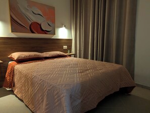 A neatly made bed with a patterned quilt, a bedside table with a lamp, a wall-mounted artwork, and a window with curtains. - Tarrafal Bay · 1Bdr Apt · Sea view - 4 (Tarrafal)