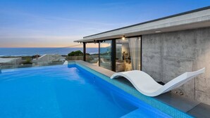 Una piscina moderna con vista despejada al océano, un área de estar techada y un muro exterior de concreto.