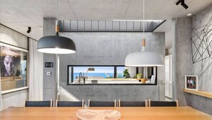 Un moderno comedor con una larga mesa de madera, lámparas colgantes blanco y negro, vista al mar a través de una ventana y una pared de concreto con una obra de arte geométrica.