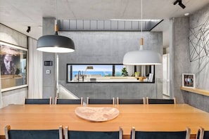 A modern dining area with a long wooden table, black and white pendant lights, a view of the sea through a window, and a concrete wall with a geometric art piece. - Luxury Coastal Escape pool | sauna | cinema| views (Ocean Grove)