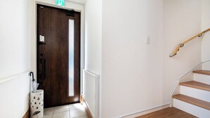 Un corridoio con una porta in legno, una parete bianca, una scala in legno e un cestino bianco con un motivo piovoso.